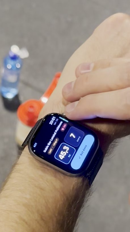 En Apple Watch på håndleddet ute på et treningssenter. Løfteren skrur inn 47 kg og 7 reps i Musklr på Cable Bar Lateral Pulldown, trykker på Set done, og klokka feirer en personlig rekord med konfetti: 47 kg, 6 kg mer enn for tre måneder siden.